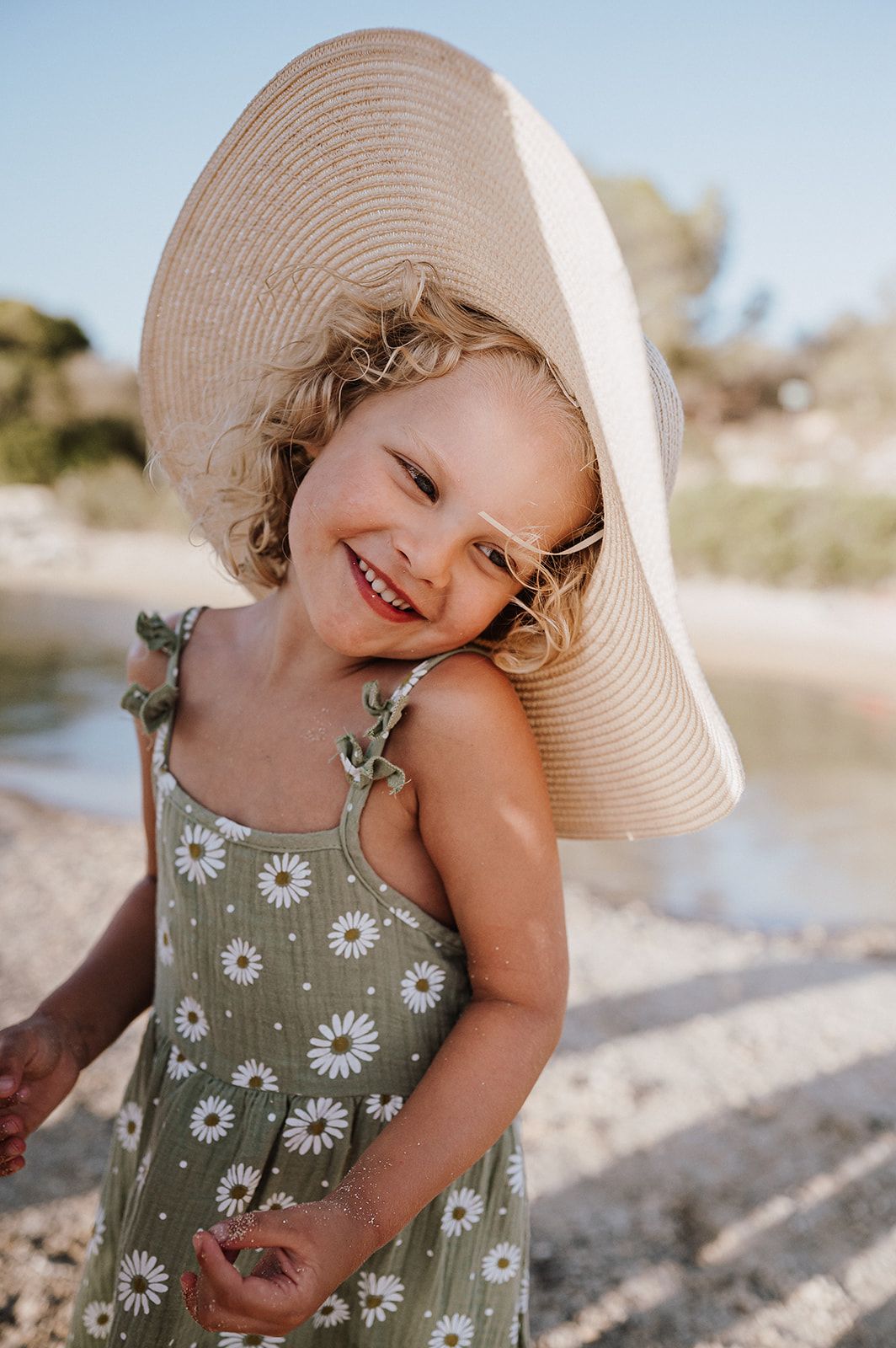Adorable curly-haired girl smiling in a daisy dress and oversized sun hat.