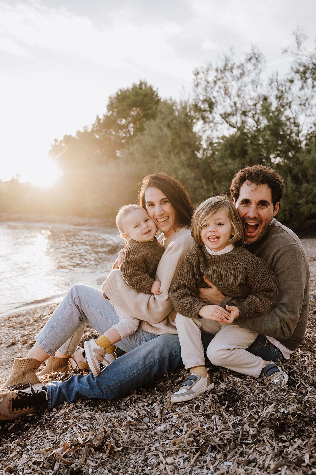 Happy family of four sitting together on a sunny beach shore, parents hugging two young children at golden hour.