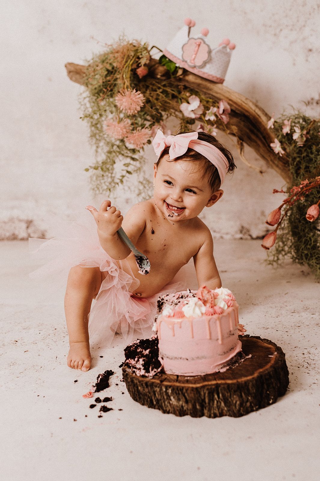 Smiling baby girl in pink tutu smashes her first birthday cake on a rustic wood slice with floral backdrop.