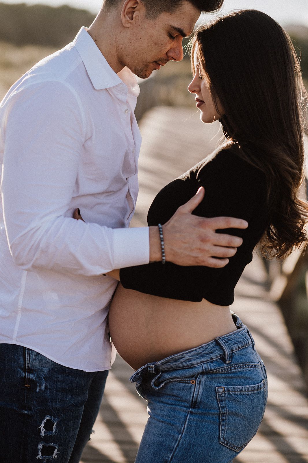 A couple shares an intimate moment outdoors, the woman visibly pregnant, wearing a black crop top and jeans.