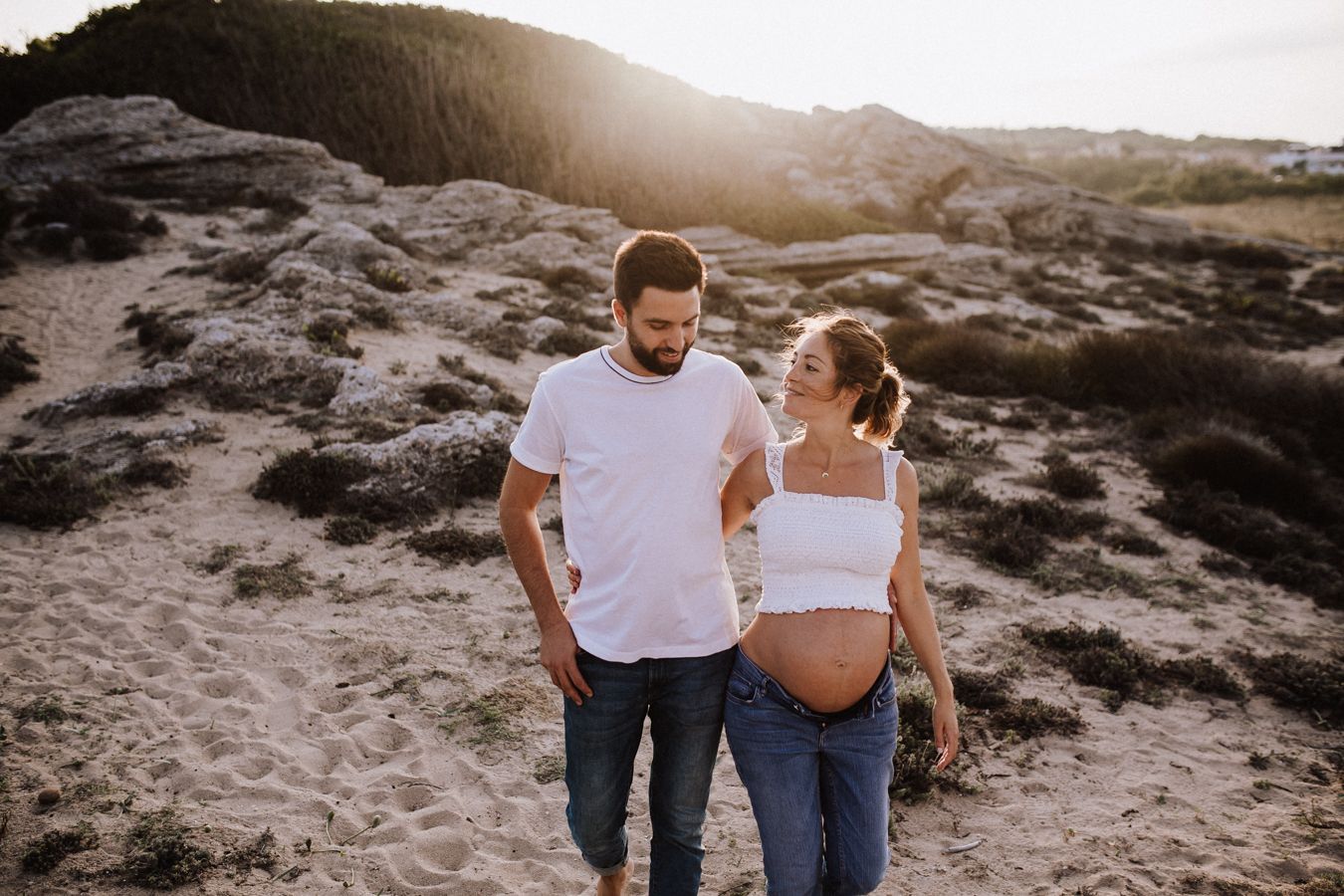 A happy couple outdoors at sunset, the woman pregnant, both wearing white tops and jeans on sandy terrain.