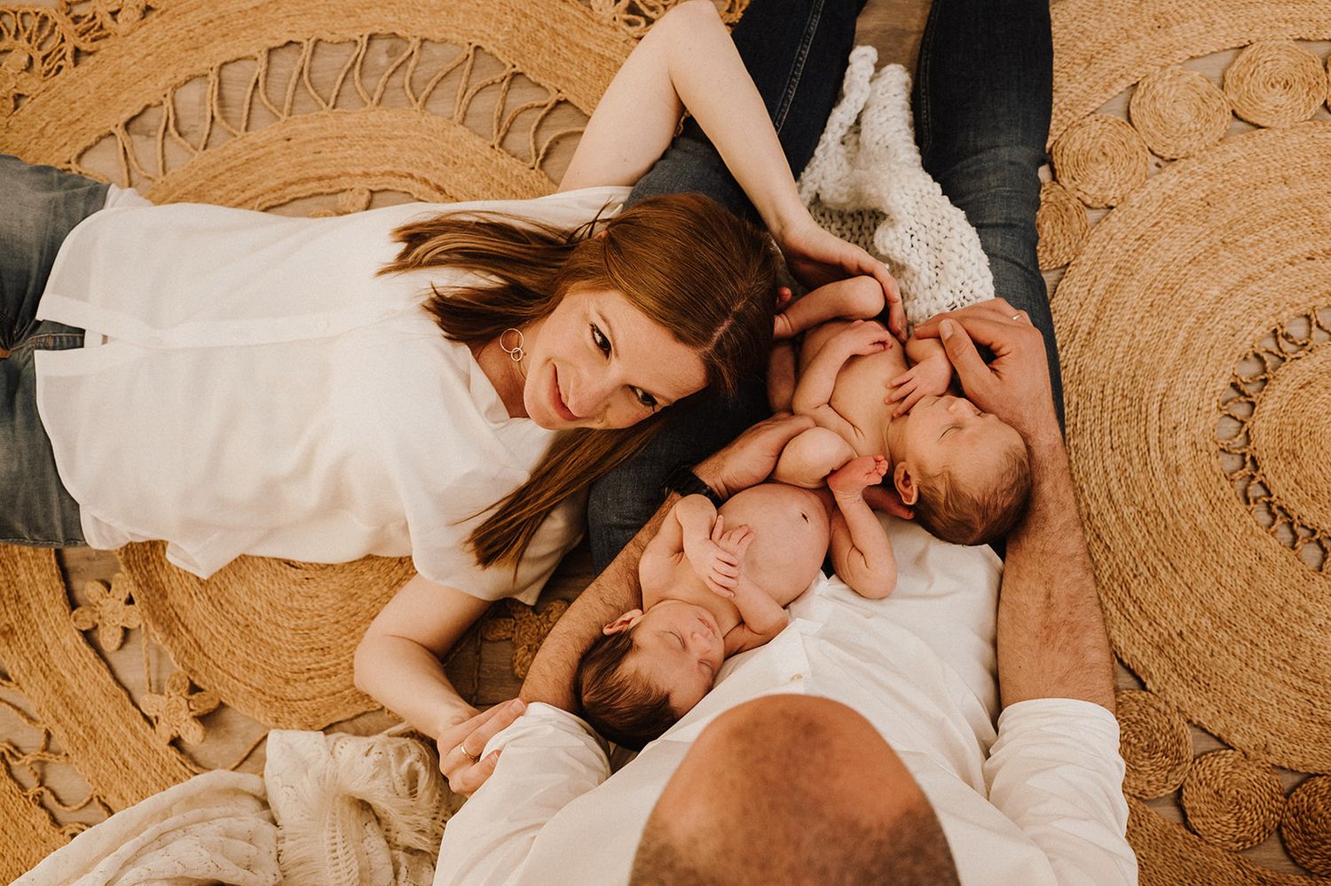 Parents lying on a woven rug, lovingly gazing at their newborn twins during a cozy family photo session.