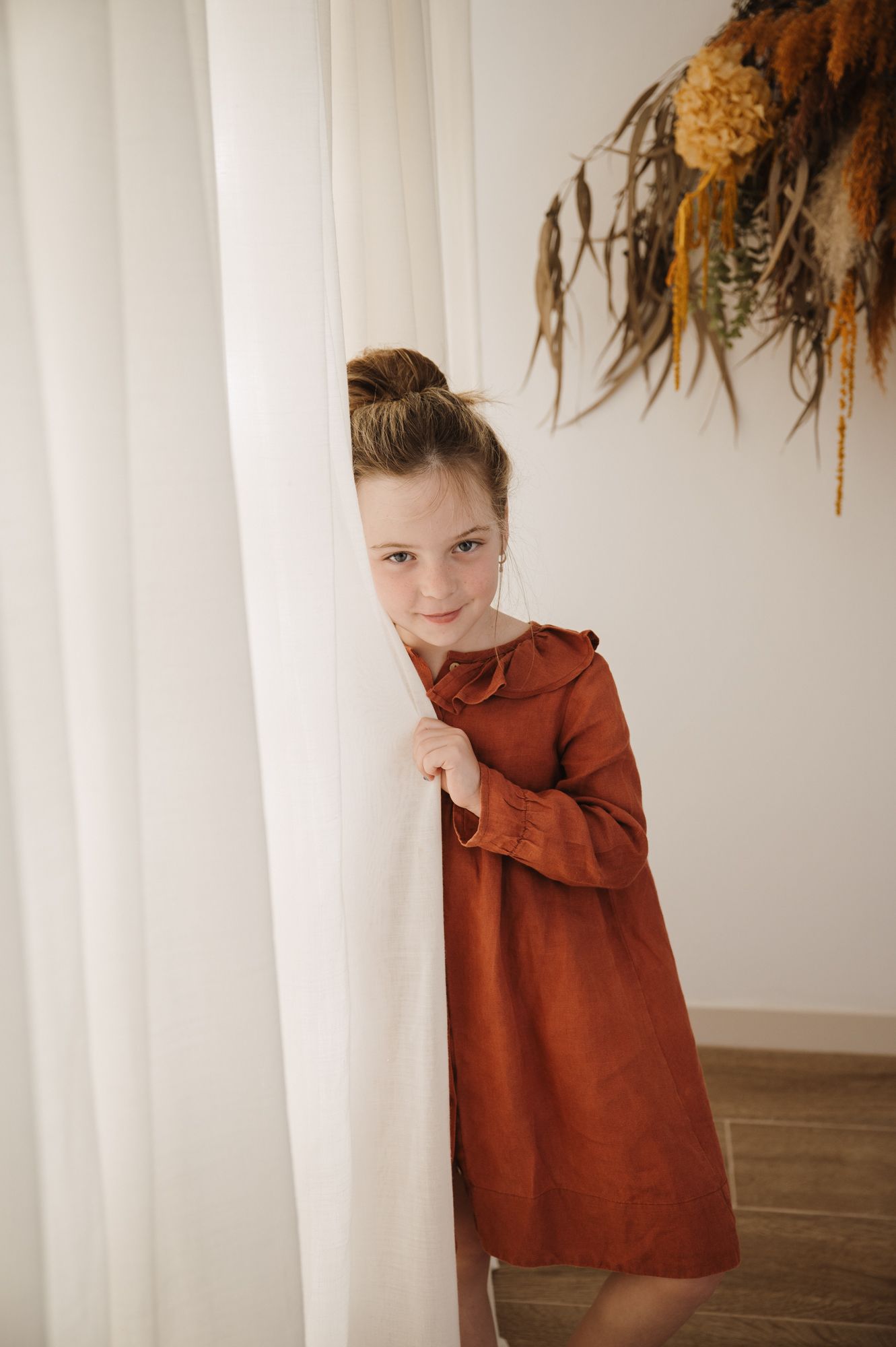 Young girl in rust-colored long sleeve dress peeking behind white curtain with dried floral arrangement above.