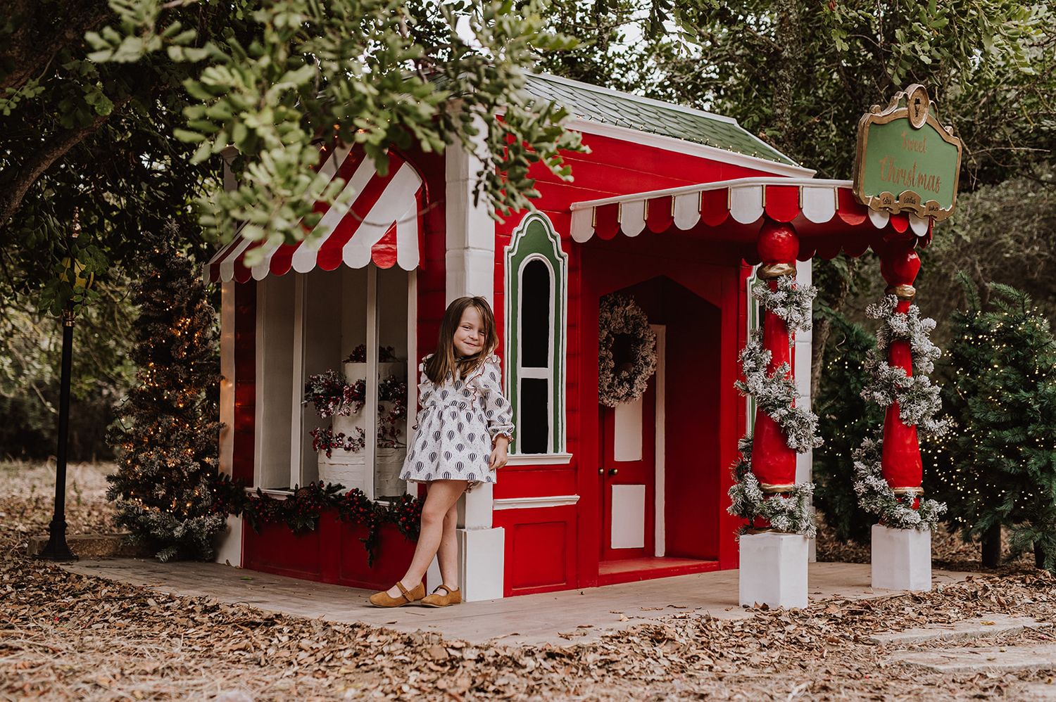 Young girl posing beside a festive red and white playhouse decorated with Christmas wreaths and garland outdoors.