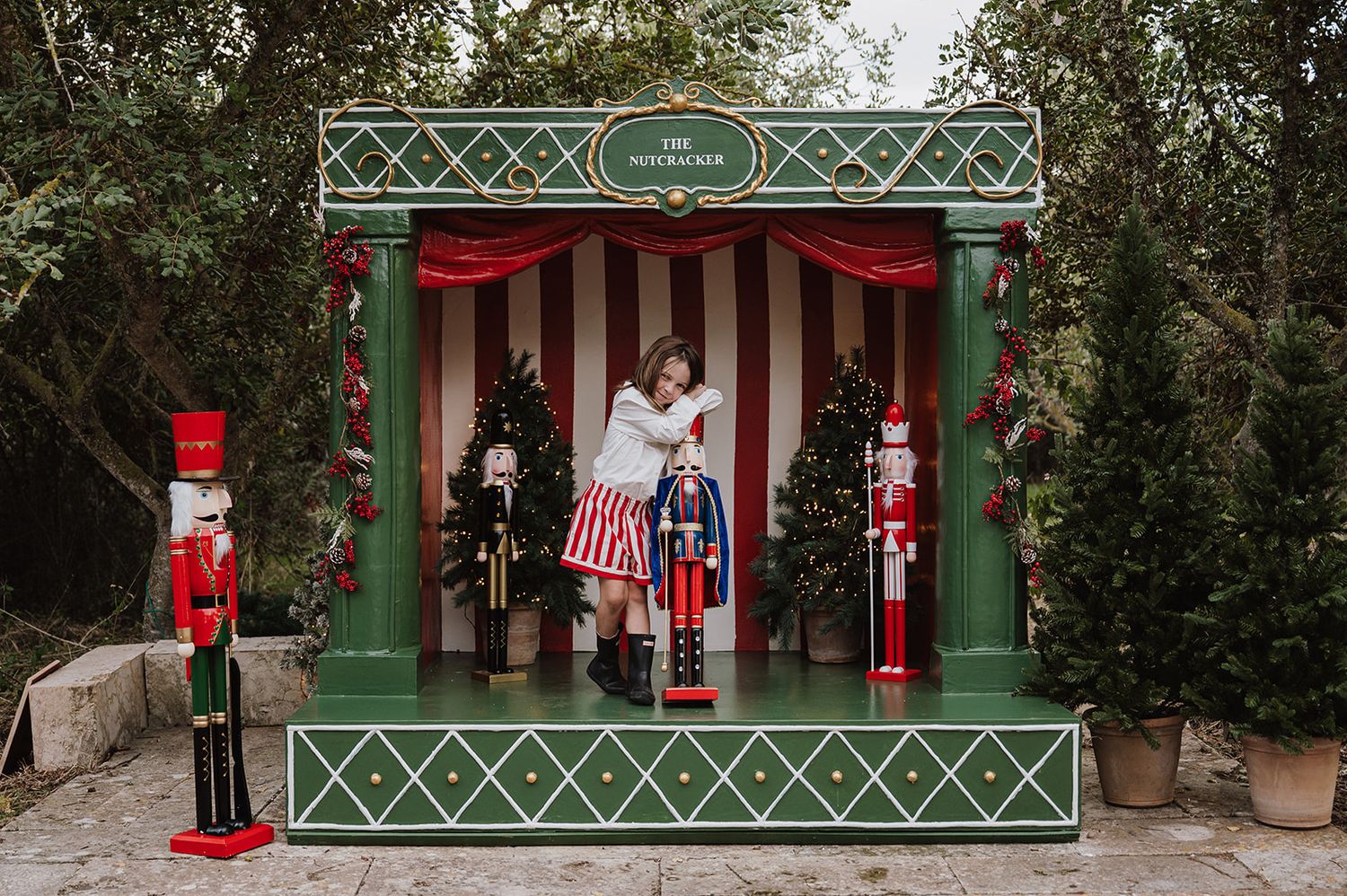 Woman poses in a festive Nutcracker-themed stage display with red striped curtains and nutcracker figurines outdoors.