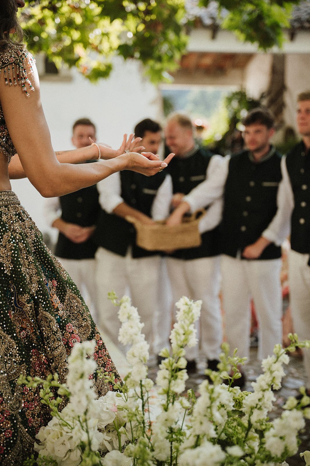 Bride in dark embroidered lehenga performing ceremony gestures before groomsmen in black jackets.