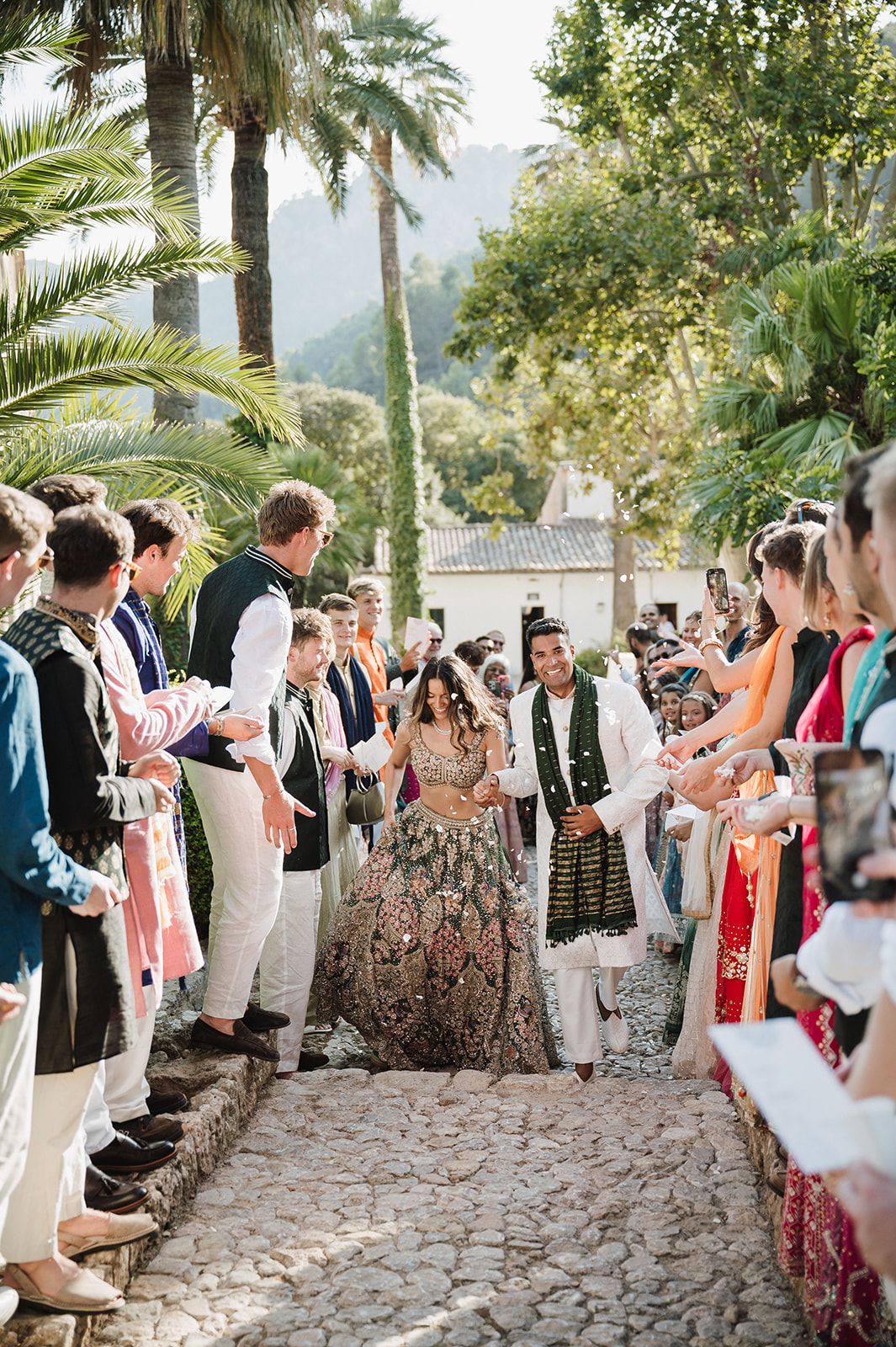 Bride and groom in Indian wedding attire walk through cheering guests at outdoor venue.