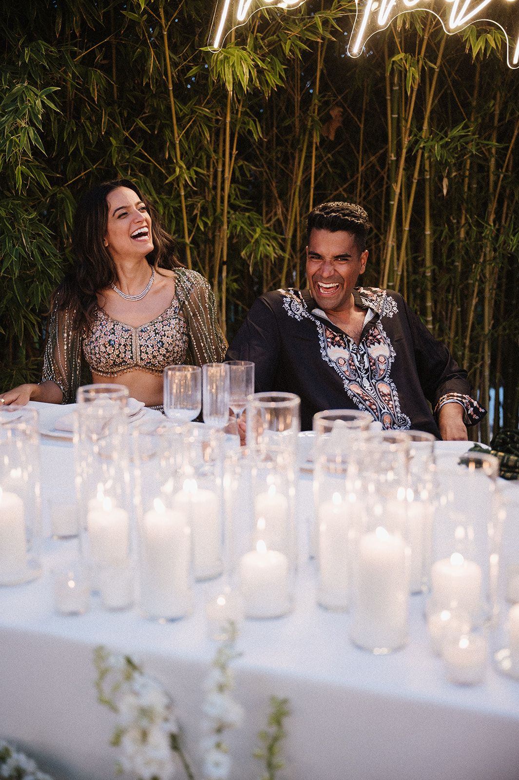 Bride and groom laugh together at candlelit wedding reception table with bamboo backdrop and neon sign.