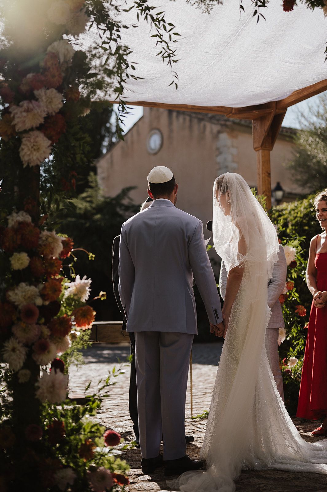 Bride in lace gown and groom in grey suit standing under a floral chuppah during an outdoor Jewish wedding ceremony.
