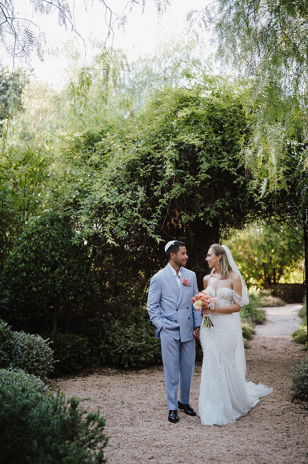 Bride and groom walk garden path surrounded by green hedges, groom in light blue suit, bride holding bouquet.