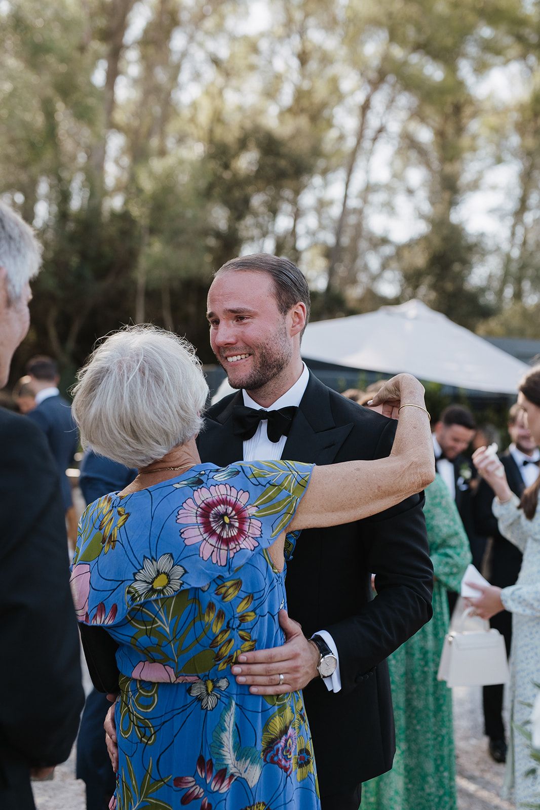 Groom in tuxedo shares a dance with older woman in floral blue dress at outdoor wedding reception.