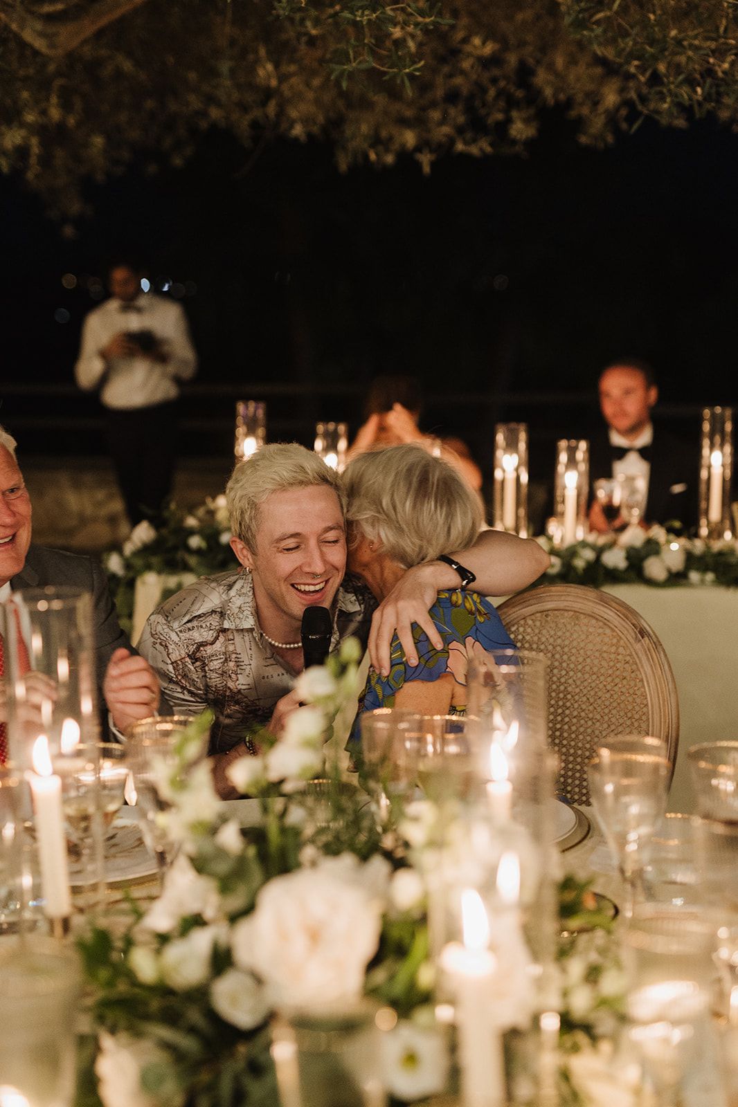 Wedding guest laughing and hugging elderly woman at candlelit reception dinner table at night.