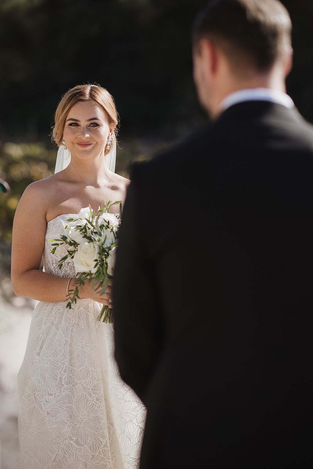 Smiling bride in strapless lace gown holding white bouquet during outdoor wedding ceremony.