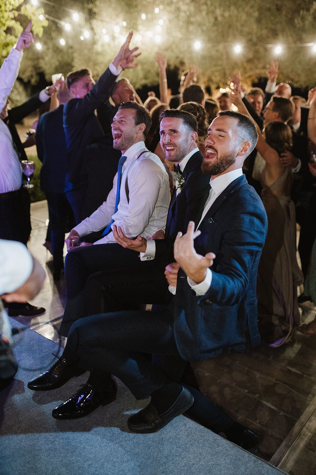 Three men in suits laugh and cheer together while seated at a lively outdoor wedding reception.