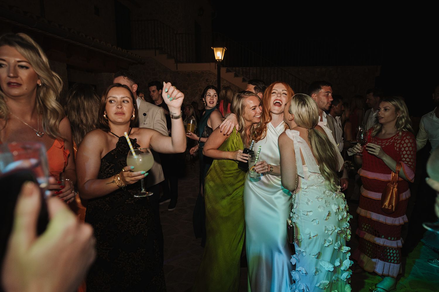Guests in colorful dresses dancing and laughing at a lively outdoor wedding reception.