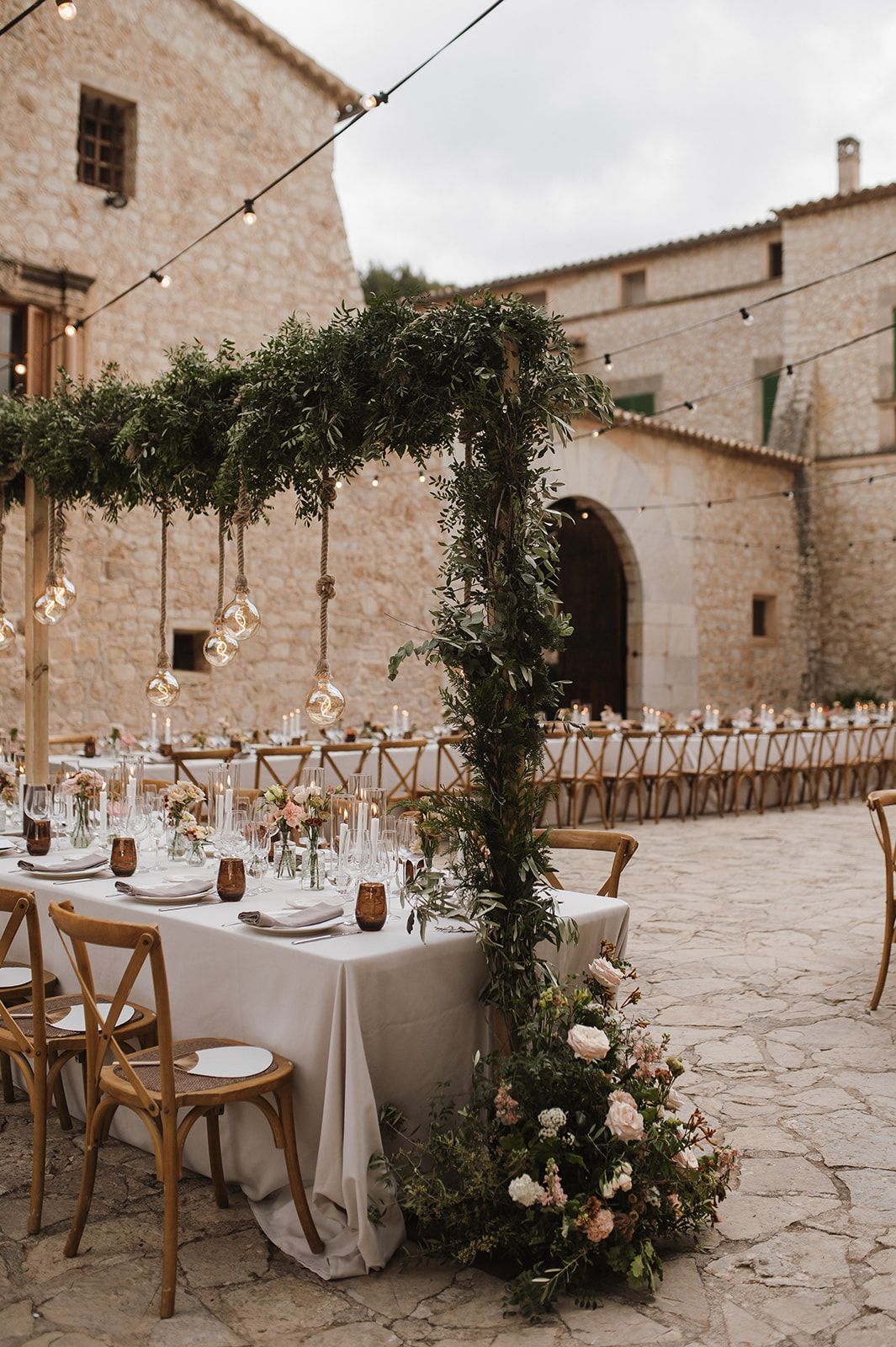 Outdoor wedding reception with lush greenery arch, white draped tables, blush floral arrangements, and cobblestone courtyard.