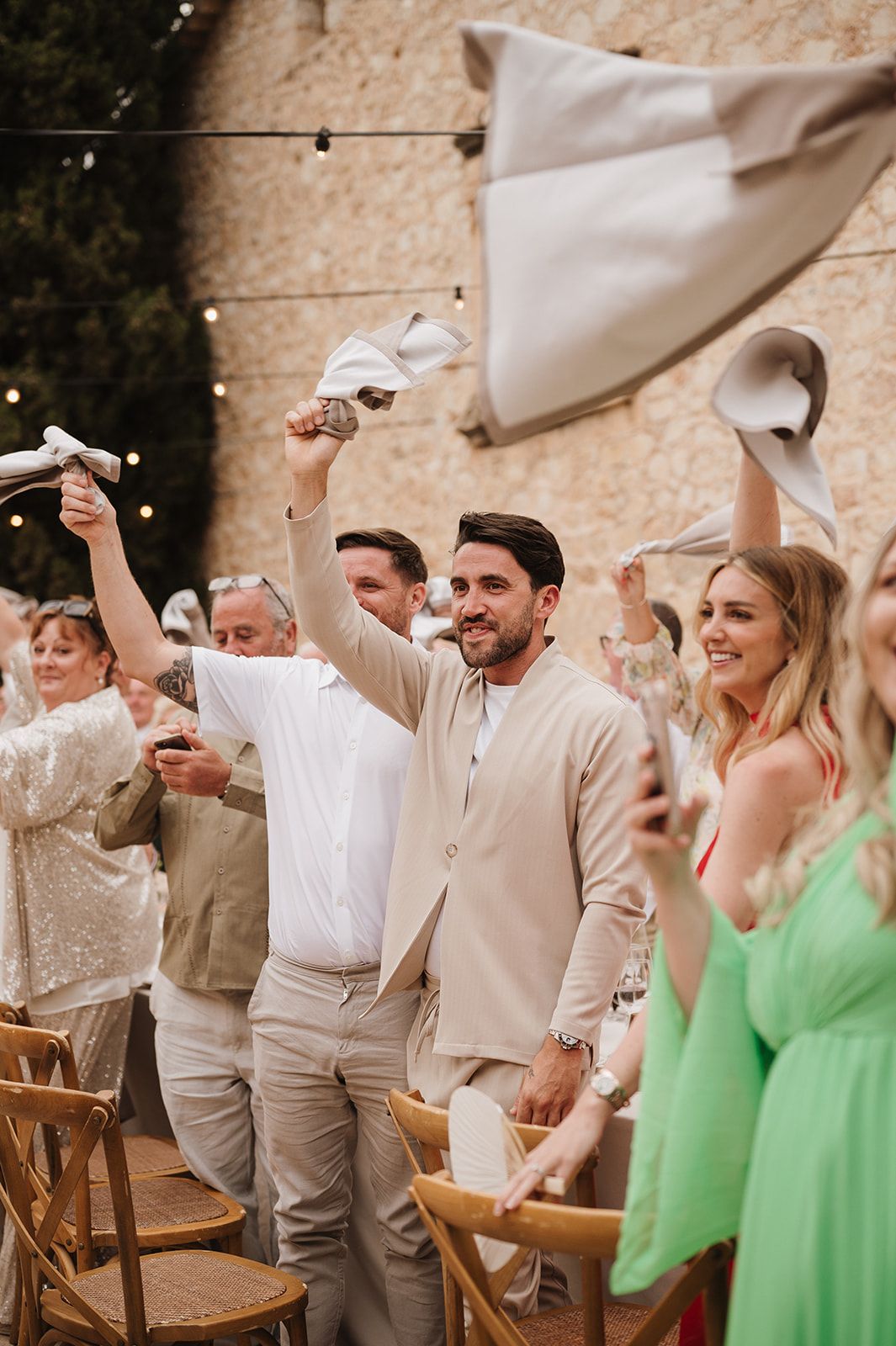 Smiling groom in beige suit waves napkin with cheering wedding guests at outdoor reception.