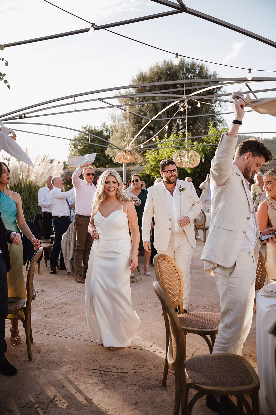 Bride and groom make grand entrance into outdoor wedding reception as guests cheer under string lights.