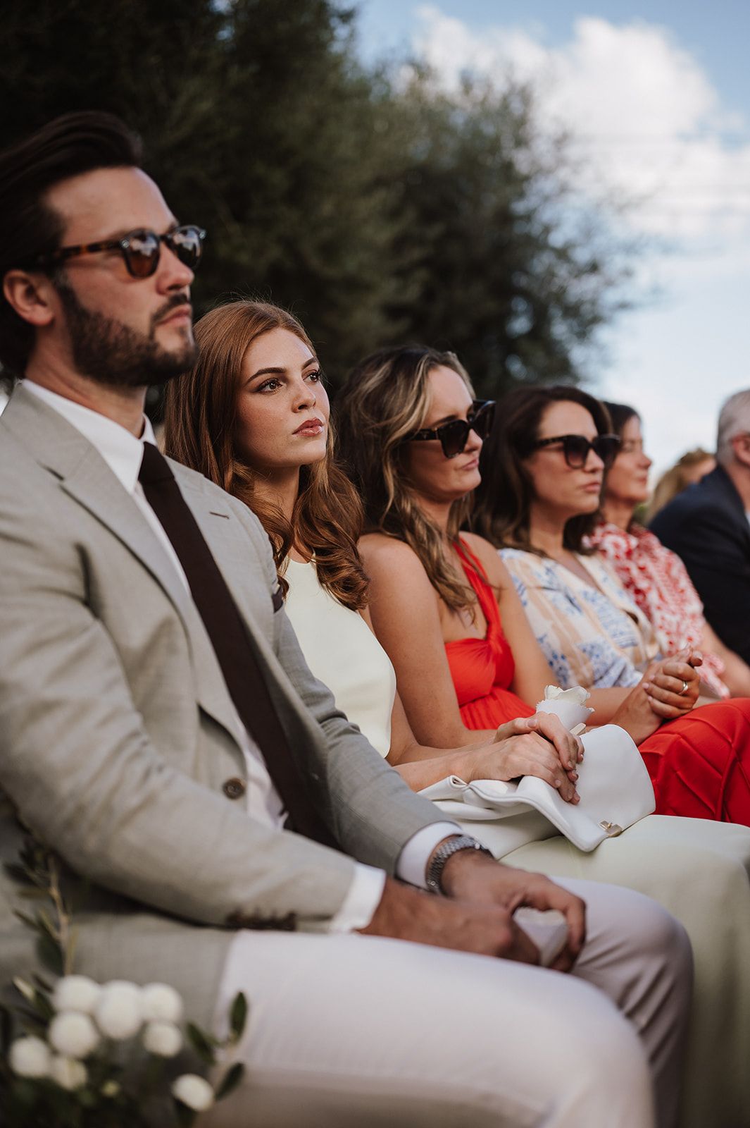 Wedding guests seated outdoors wearing sunglasses, including woman in red dress and man in beige suit.