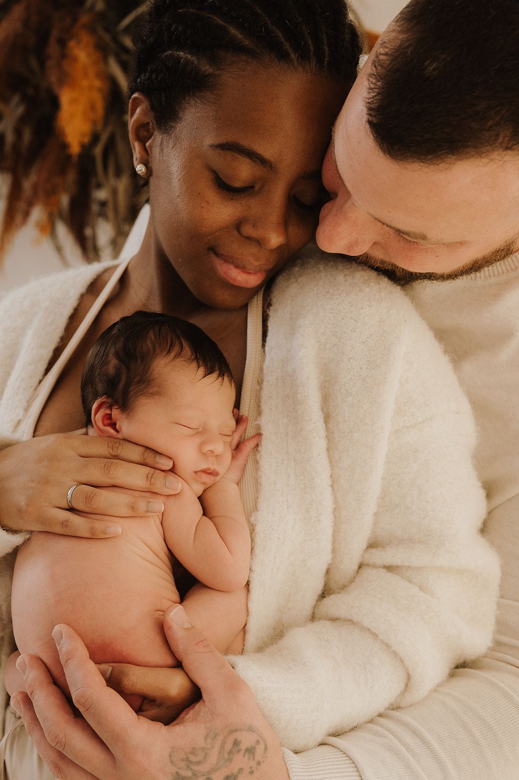 Interracial couple tenderly holding their sleeping newborn baby, dressed in matching cream outfits.