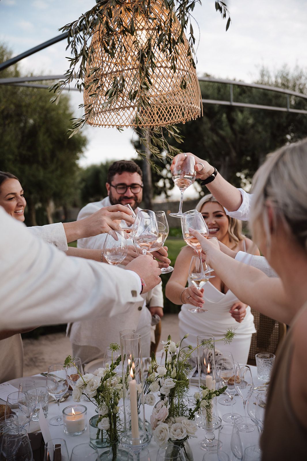 Wedding party raising champagne glasses in toast under rattan pendant light at outdoor reception.