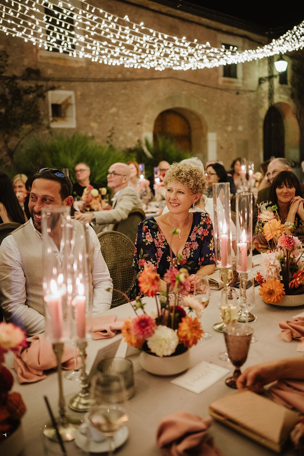 Wedding guests smile and chat at candlelit outdoor reception table adorned with colorful flowers and string lights.