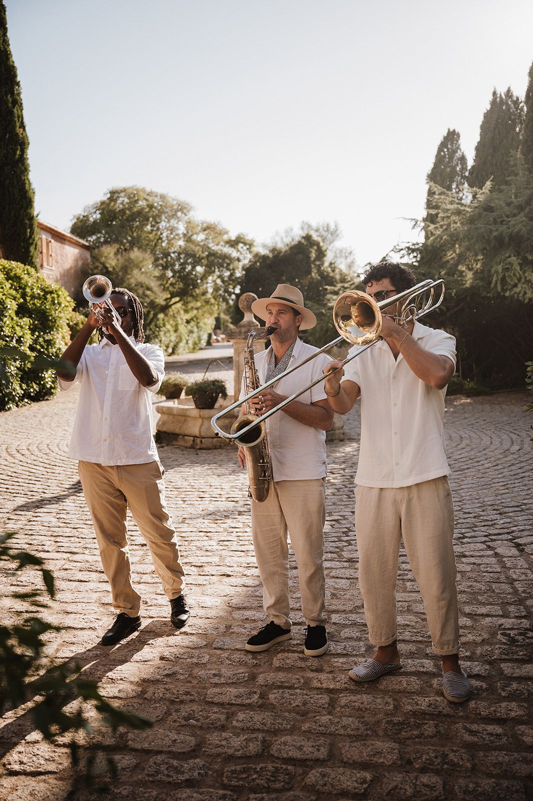 Three musicians in white shirts play brass instruments on a cobblestone path surrounded by lush greenery.