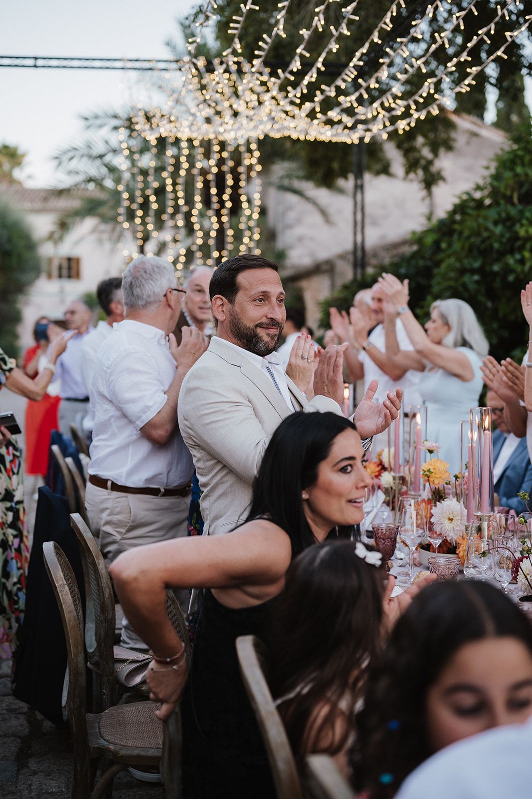 Wedding guests clap and celebrate at outdoor reception dinner under string fairy lights.