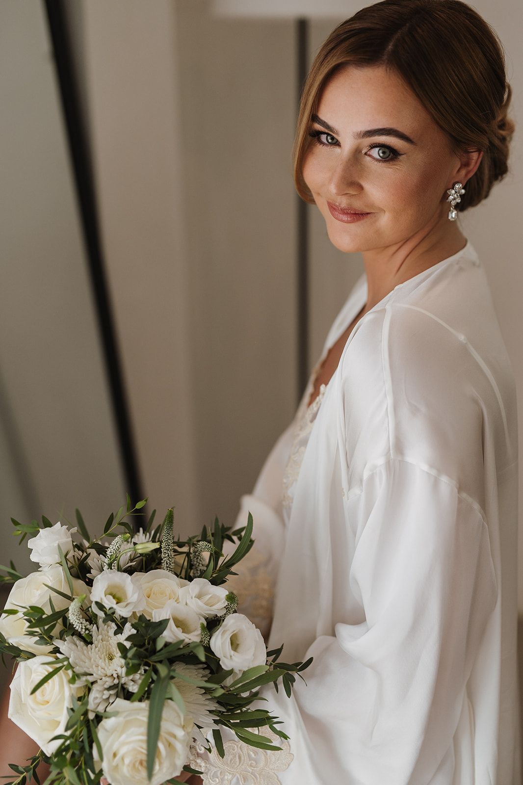 Bride in white robe holding elegant white rose bouquet with greenery, smiling softly.