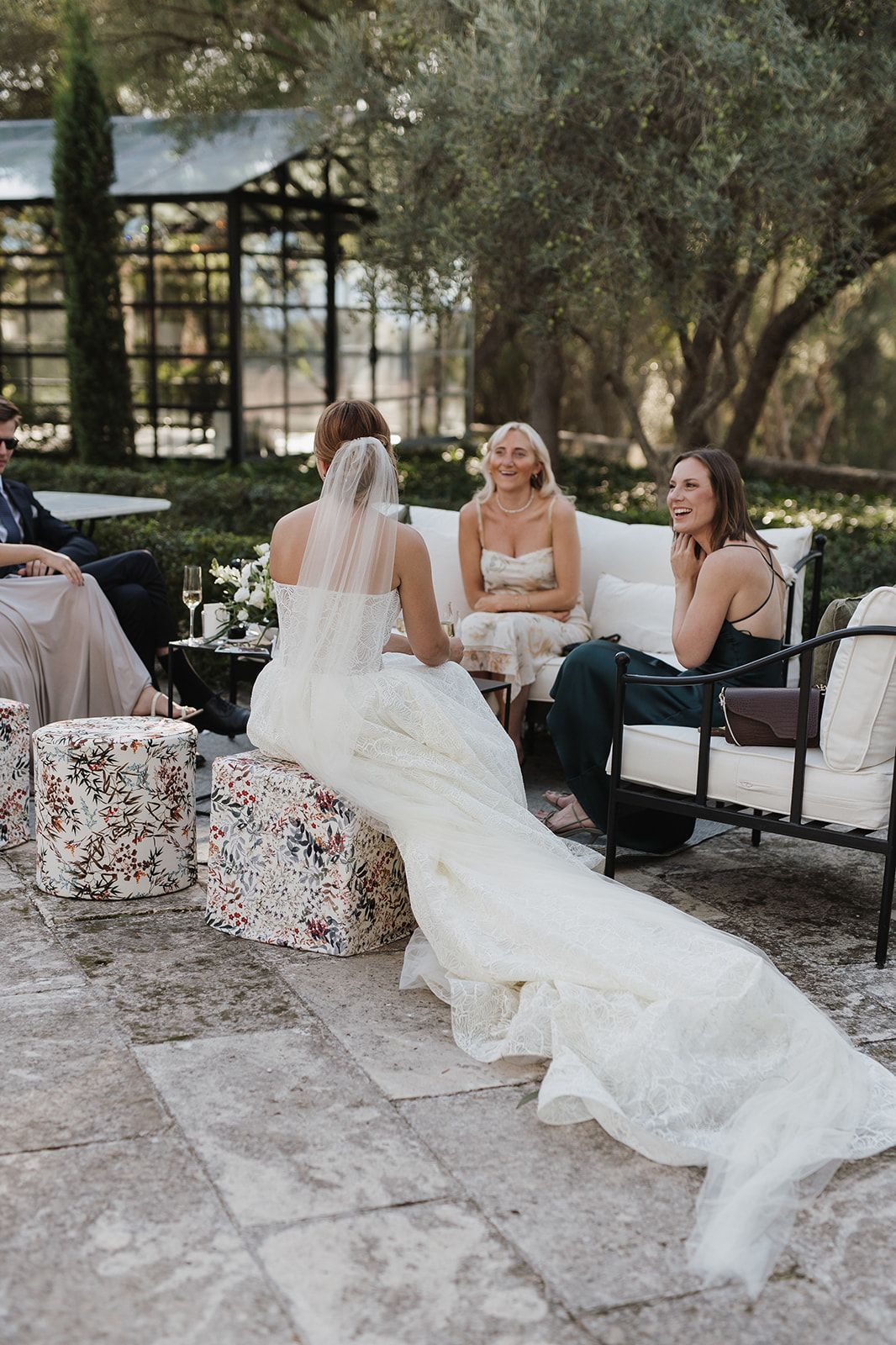 Bride in lace gown with veil sits chatting with smiling guests on patio furniture at outdoor reception.