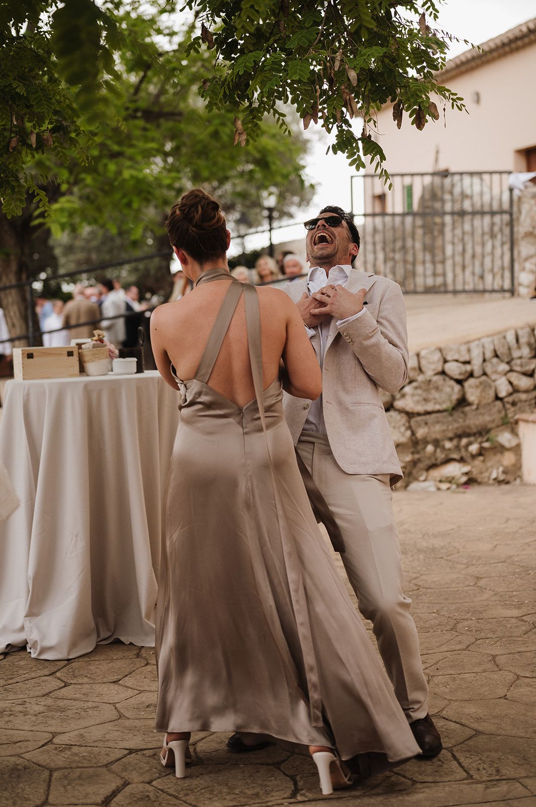 Couple in elegant formalwear dancing and laughing together at an outdoor wedding reception on stone patio.