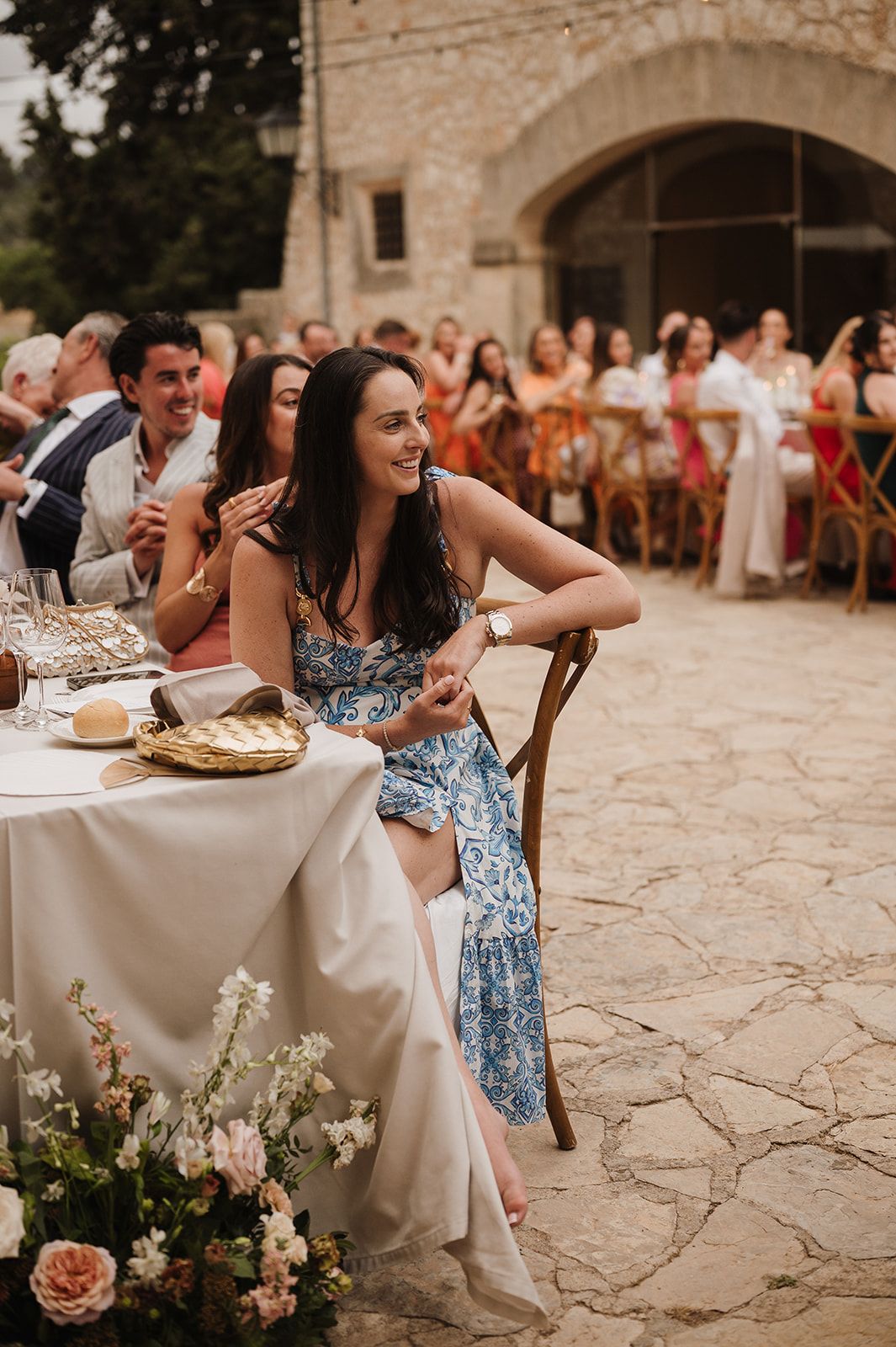Smiling wedding guest in blue floral dress relaxes at outdoor reception table with fresh flowers.