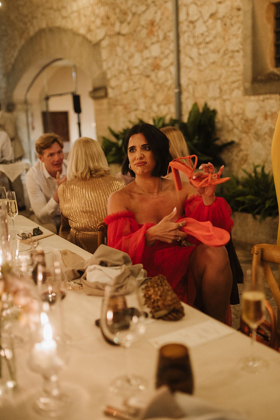 Woman in red off-shoulder dress holding pink shoes at an elegant candlelit wedding reception dinner.