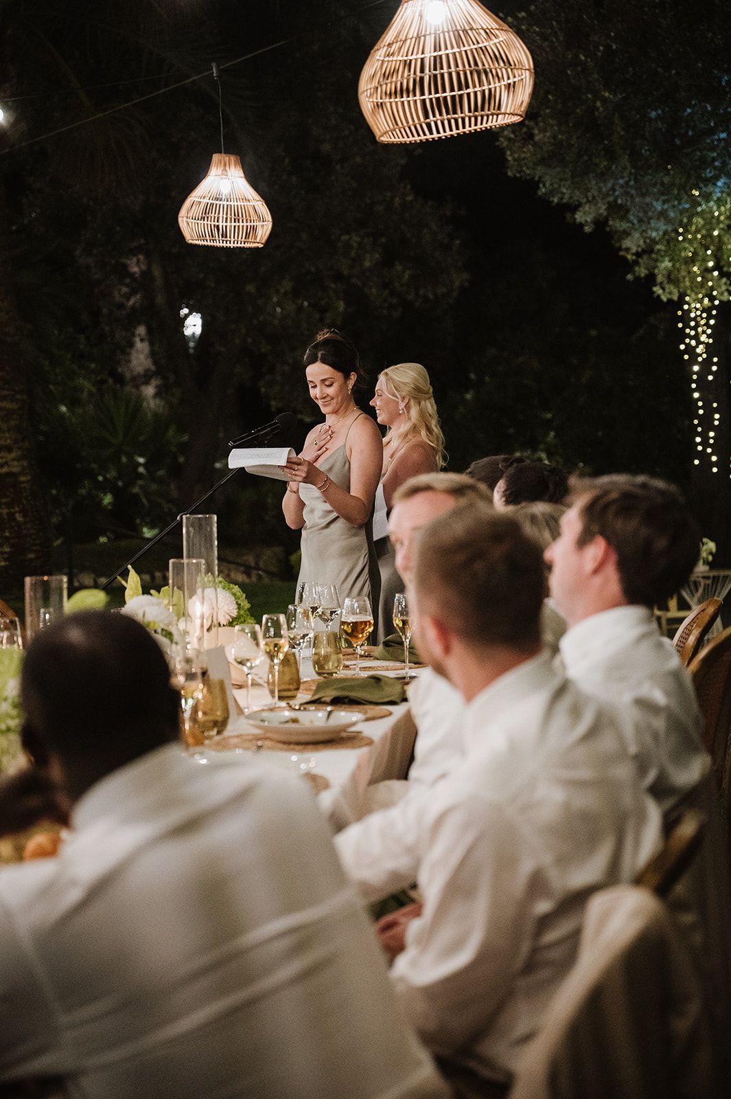 Bridesmaid delivers heartfelt speech at candlelit outdoor wedding reception beneath woven rattan pendant lights.