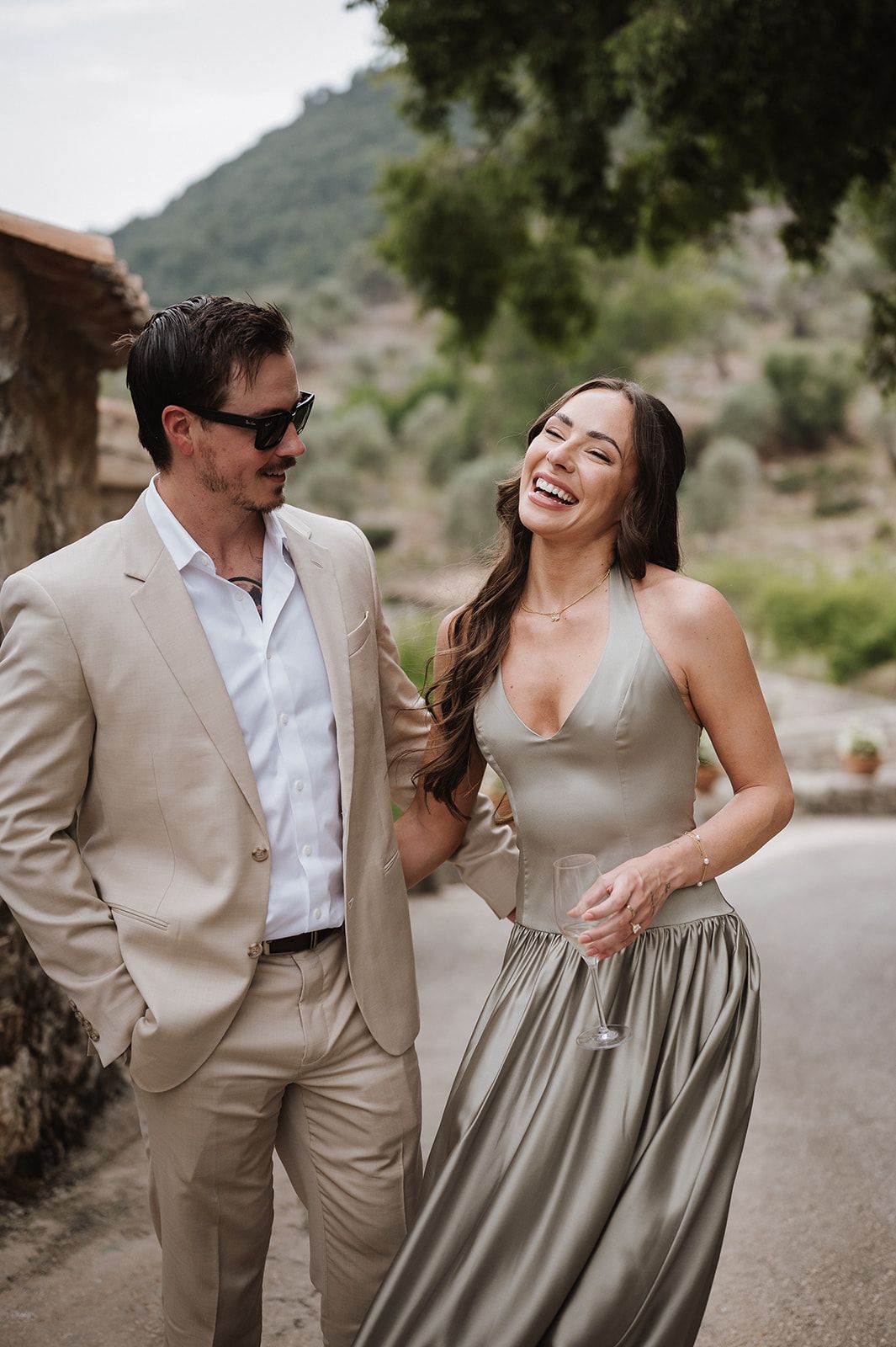 Couple laughing and walking together outdoors, man in beige suit and woman in sage green halter dress with mountains behind.