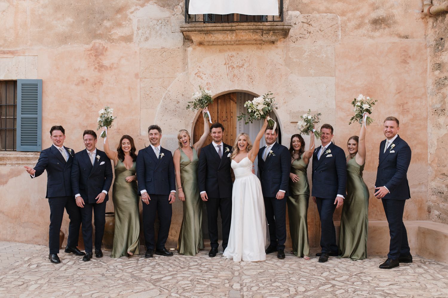 Wedding party posing in front of rustic stone wall, bridesmaids in sage green gowns, groomsmen in navy suits.
