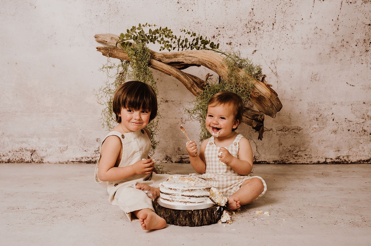 Two young children sit together on a wooden slice with a cake, posed before a rustic driftwood and greenery backdrop.
