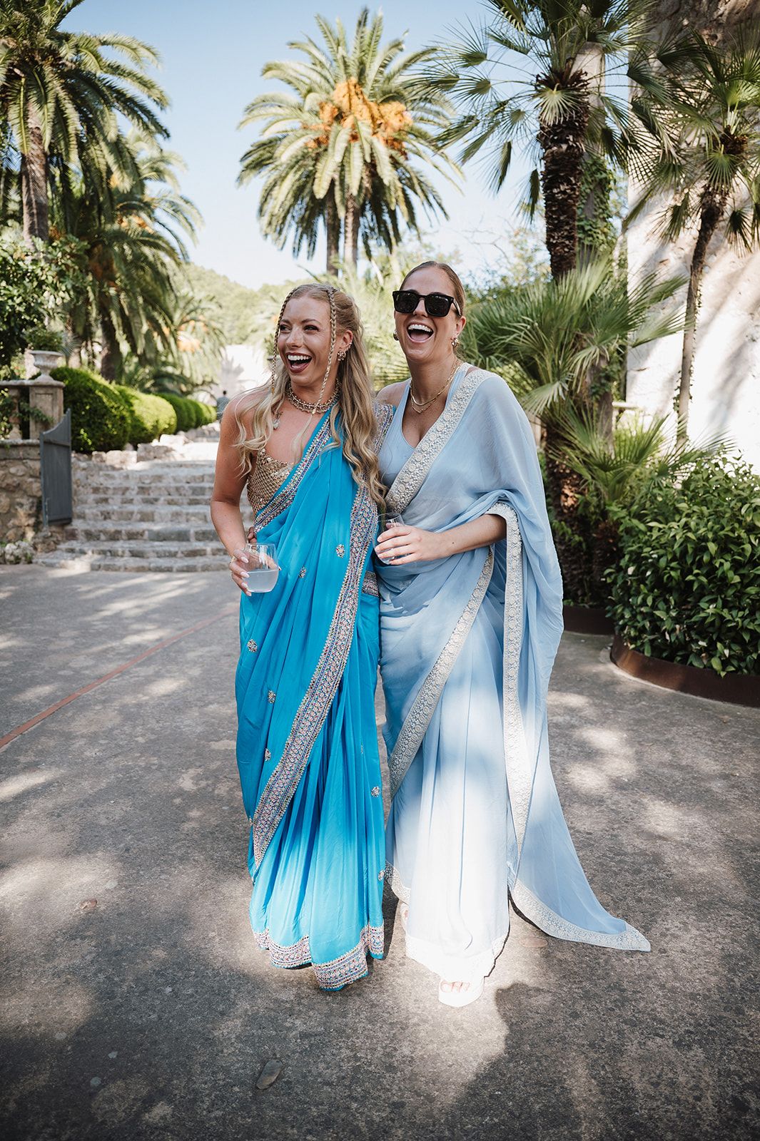 Two women laughing outdoors in blue and light blue Indian sarees with silver embroidery trim.