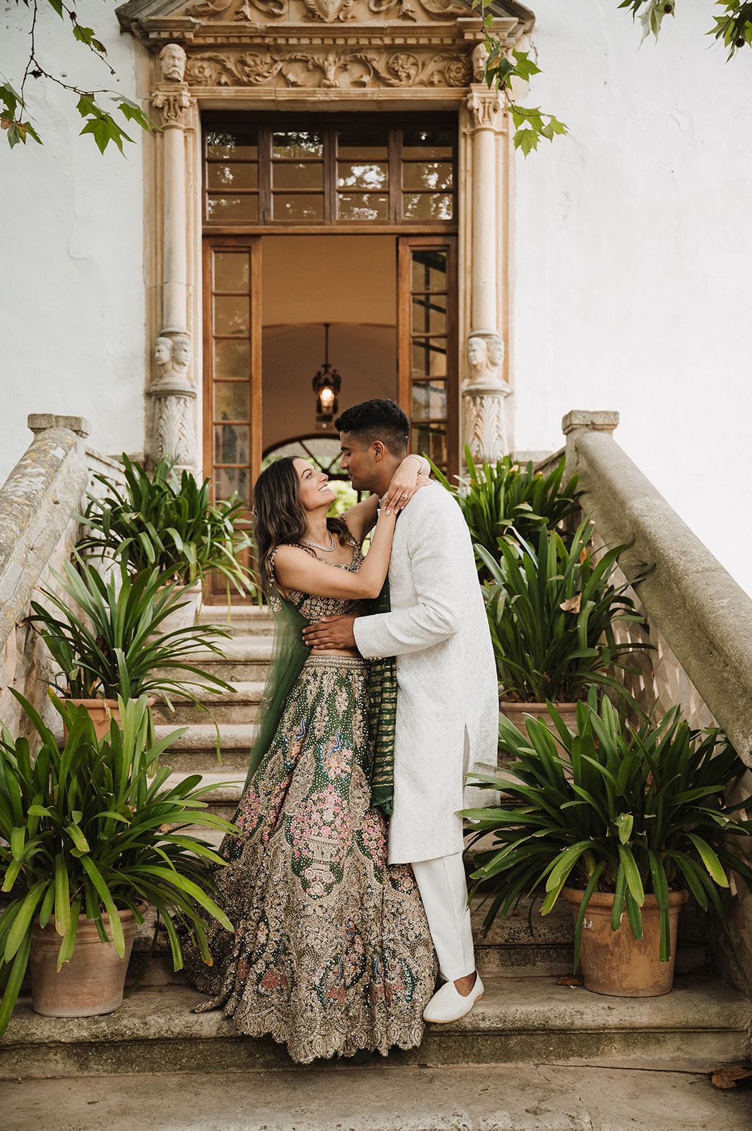 Indian wedding couple sharing romantic kiss on ornate stairway surrounded by tropical plants.