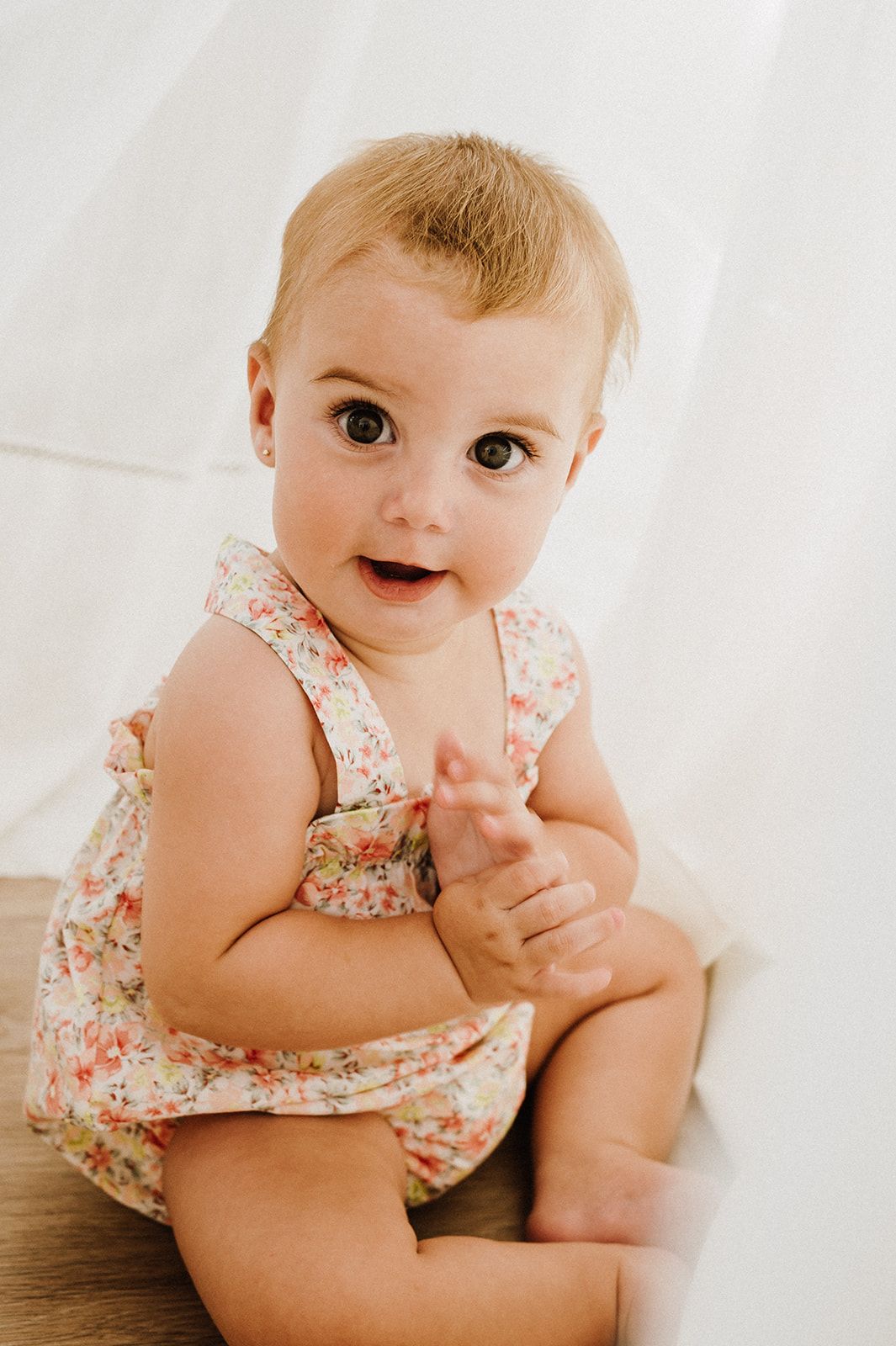 Adorable baby girl in floral romper sitting on wooden floor, looking at camera with big brown eyes.