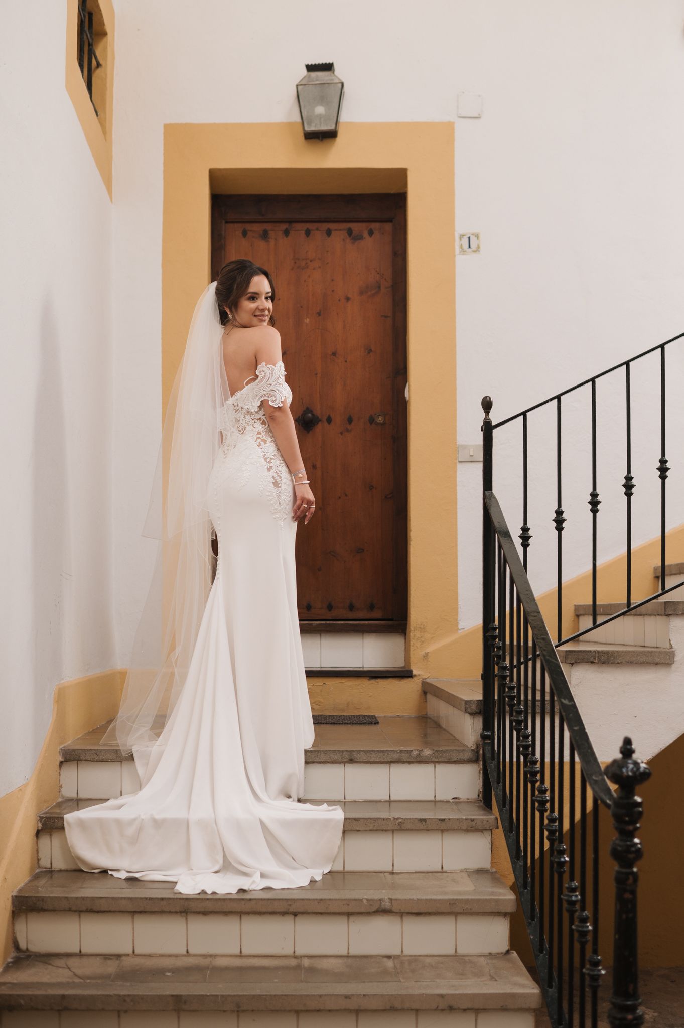 Elegant bride in a white off-shoulder lace wedding gown with veil stands on stone steps before a wooden door.