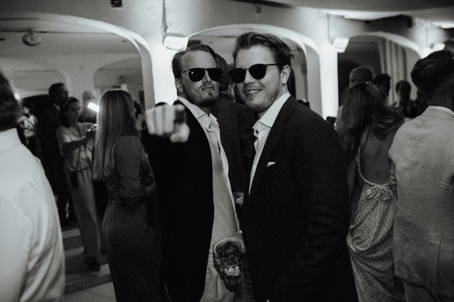 Two men in suits wearing sunglasses pose together smiling at a wedding reception party.
