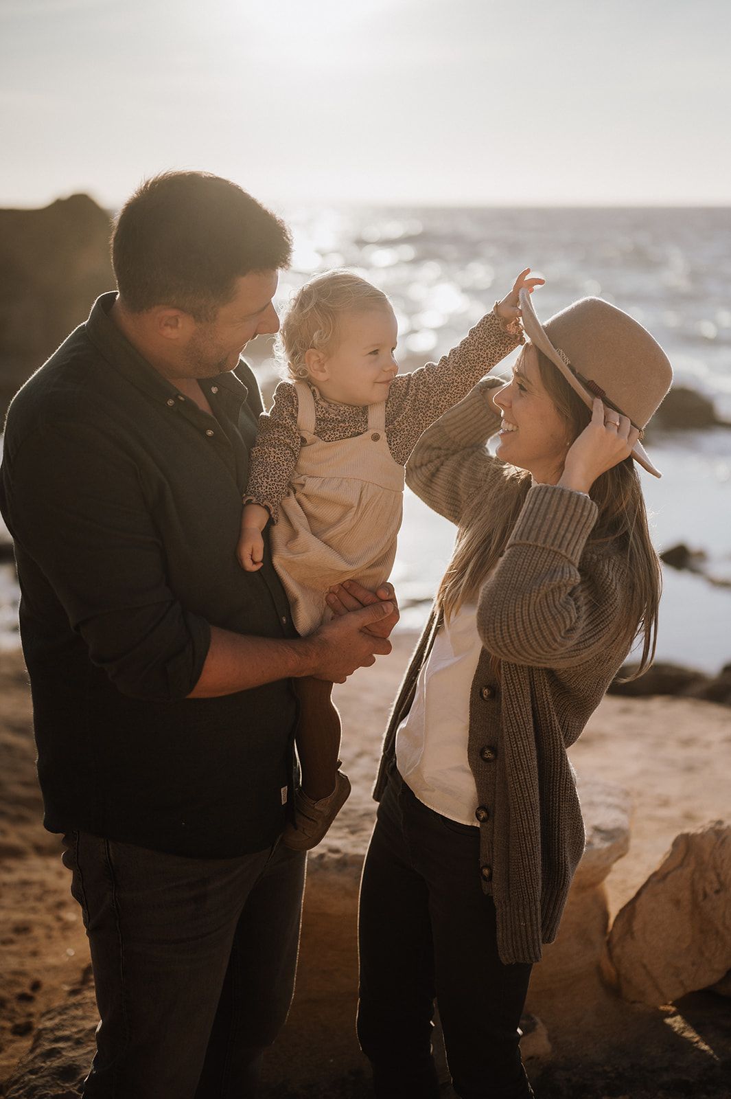 Parents interact lovingly with their baby on a sunlit rocky beach at sunset.