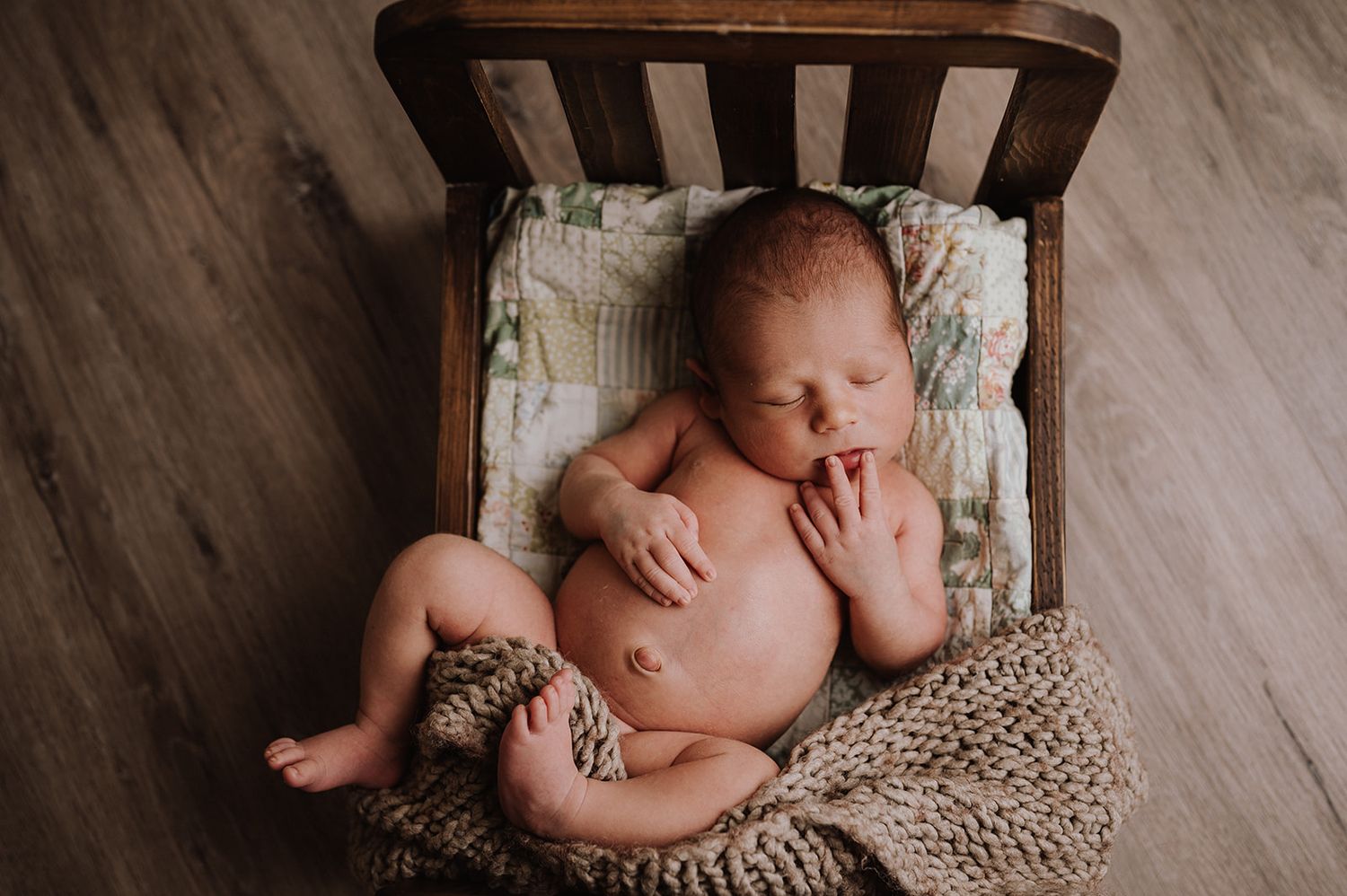 Peaceful newborn baby sleeping in a tiny wooden bed with a floral blanket and knit wrap.