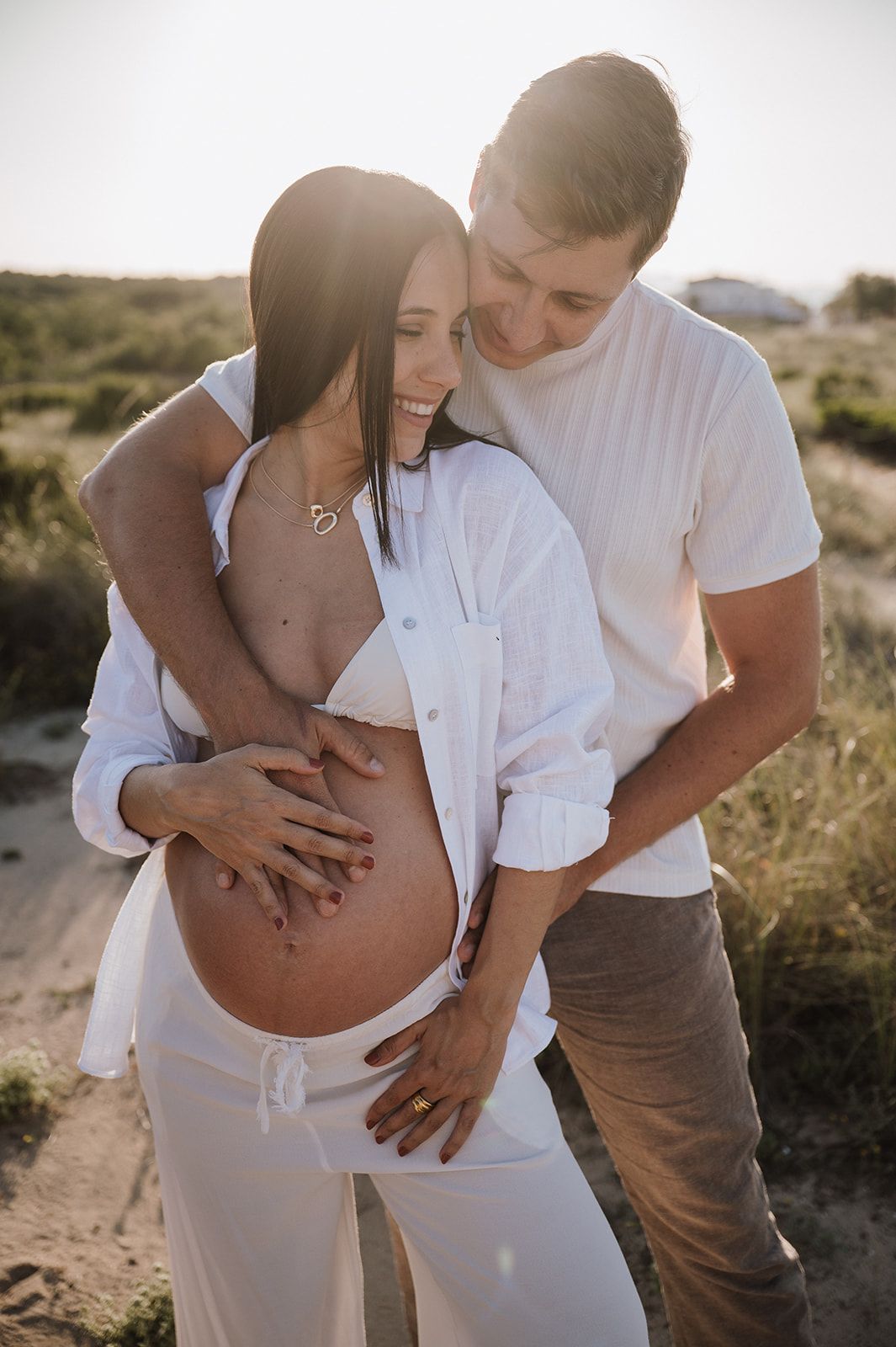 Happy couple embracing outdoors, hands on pregnant belly, smiling in warm sunset light.