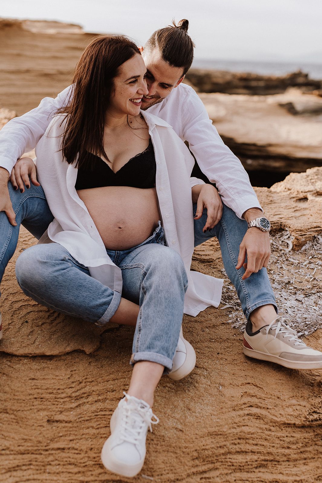 Pregnant couple sitting on sandy rocks, woman in black bralette and open white shirt showing baby bump, man in white shirt.