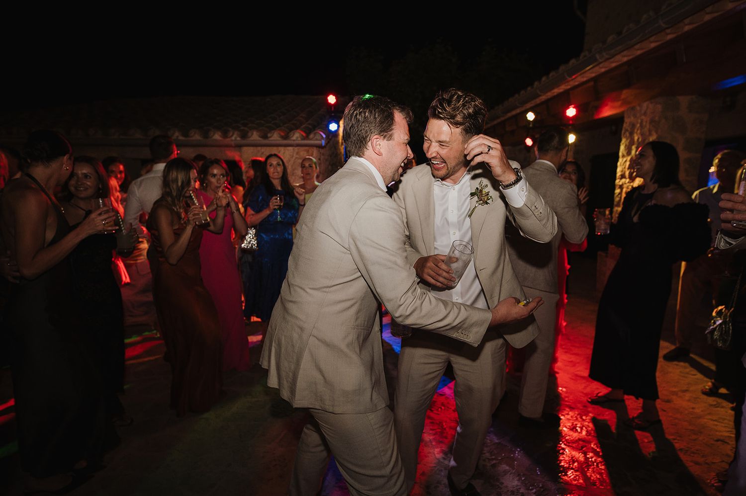 Two men in beige suits dancing together at a vibrant outdoor wedding reception at night.