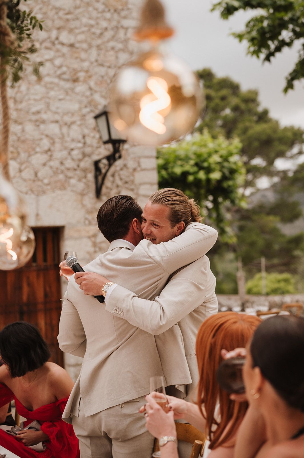 Two men in beige suits sharing a warm embrace after emotional wedding reception speeches outdoors.