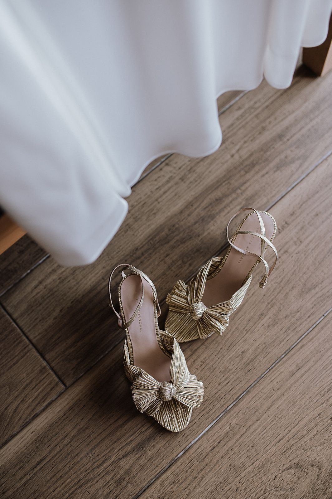Gold pleated bow heeled sandals on wooden floor beside white wedding dress hem.