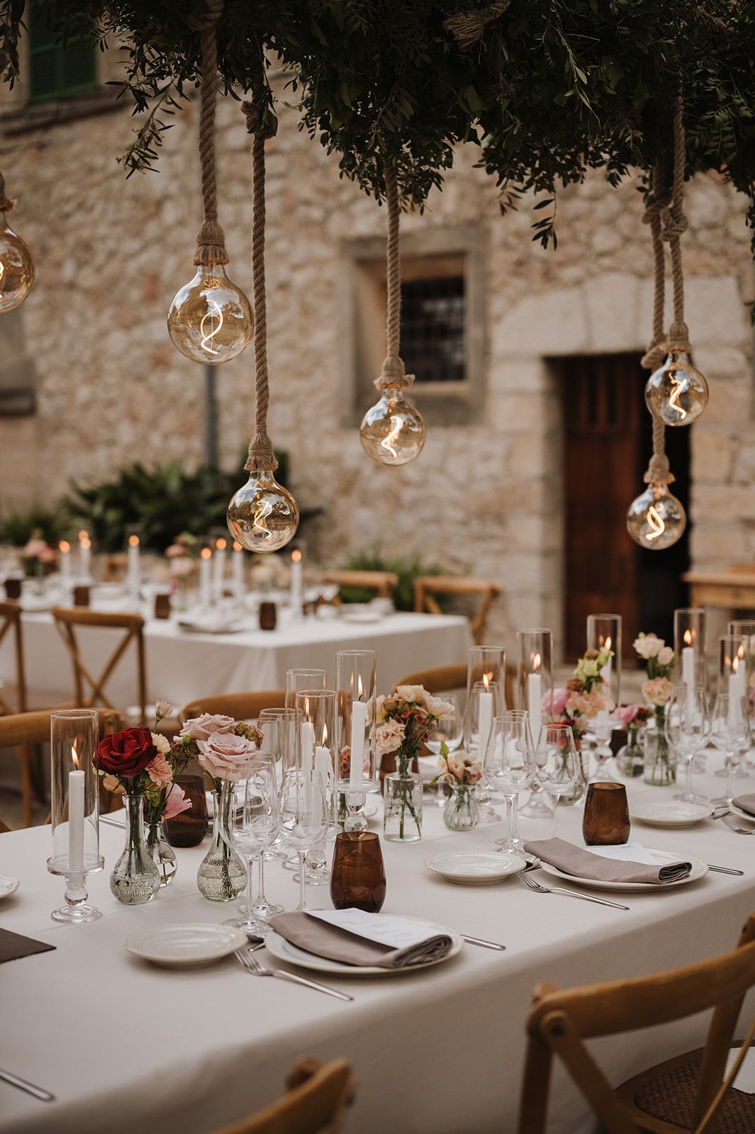 Close-up of elegant wedding table with hanging rope bulb lights, pink flowers, candles, and crystal glassware.